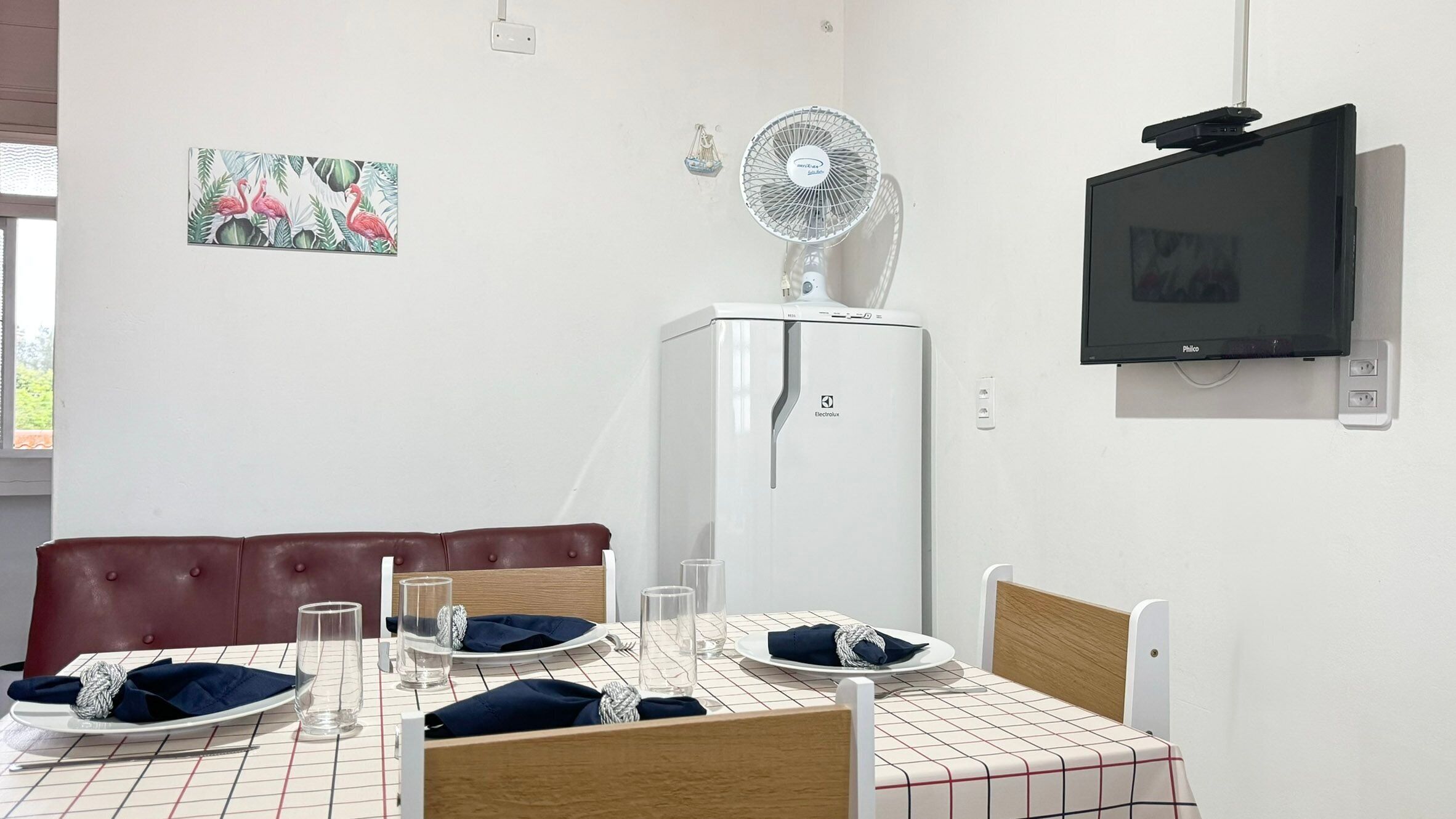 Zona pranzo con tavolo apparecchiato per due, un frigorifero bianco, una ventola a parete e un televisore.