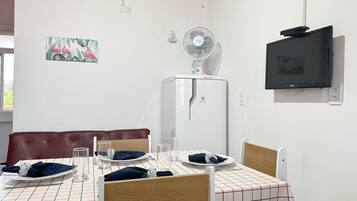 Zona pranzo con tavolo apparecchiato per due, un frigorifero bianco, una ventola a parete e un televisore.