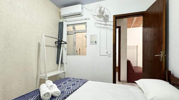Un dormitorio con cama, aire acondicionado blanco, ventana con cortinas, puerta de madera y un espejo.