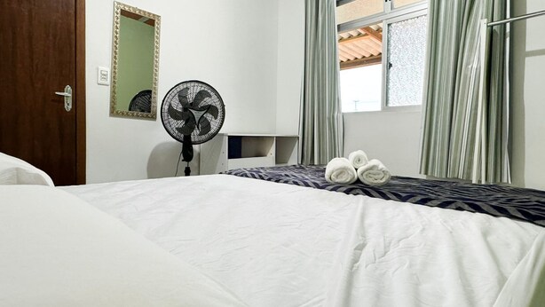 A hotel room with a bed, a standing fan, a mirror, and a window with curtains.