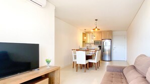 Una sala de estar moderna con televisor de pantalla plana, un mueble de madera para entretenimiento, un sofá color beige y un comedor con mesa y sillas.