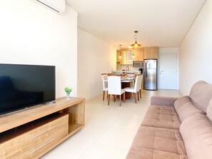 A modern living room with a flat-screen TV, a wooden entertainment unit, a beige sofa, and a dining area with a table and chairs.