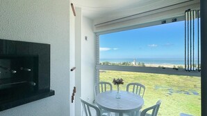 Un balcón con una mesa redonda y sillas, un televisor montado en la pared, y vista al mar.