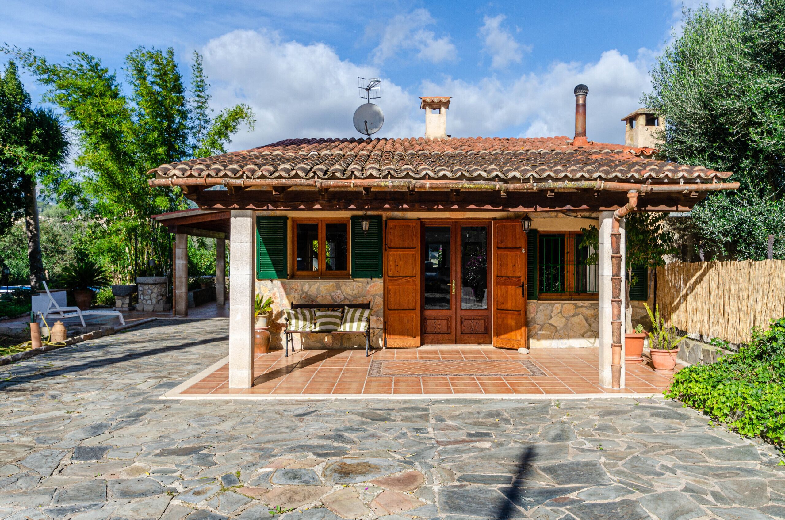 A stone-paved patio leading to a traditional house with a tiled roof, wooden doors, and a stone bench.