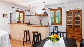 A kitchen with a central island, wooden chairs, a dining table with a vase of fruit, and a cabinet with glass doors.