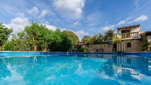 A clear swimming pool surrounded by lush greenery and a stone building with a balcony.