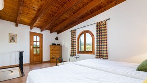 A bedroom with two beds, wooden ceiling beams, a wooden dresser, a door with a window, and a view of the outdoors.
