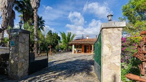 A stone pathway leading to a rustic building with a tiled roof, surrounded by lush greenery and palm trees.