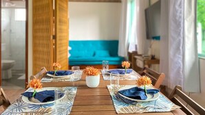 A dining area with a wooden table set for four, blue napkins, and orange flowers. A blue sofa is in the background, and a TV is mounted on the wall. - Casa Recanto da Armação - SDI001 (Florianópolis)