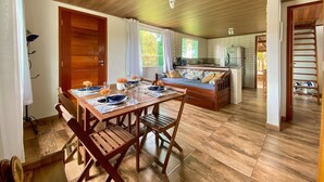 A cozy, open-plan living space with a wooden dining table set for four, a sofa, and a kitchen area. - Casa Recanto da Armação - SDI001 (Florianópolis)
