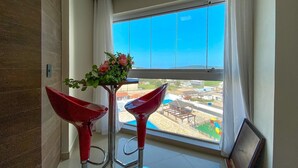 Una habitación con un ventanal grande, un taburete rojo y un florero con flores.