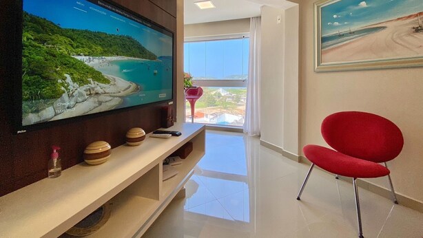Una sala moderna con un televisor de pantalla plana grande sobre un mueble bajo de madera, una silla roja y un cuadro de una escena de playa en la pared.
