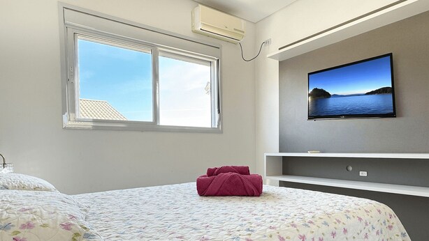 A bedroom with a bed, a window, a TV, and a wall-mounted shelf.