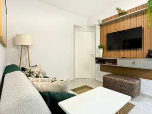A modern living room with a white sofa, a coffee table, a TV mounted on a wooden panel wall, a lamp, and a small plant.