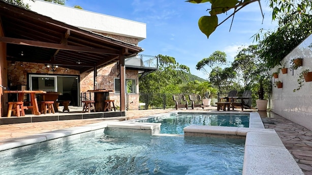 A pool area with stone-tiled steps, wooden lounge chairs, and a covered seating area with tables and chairs. - Paraíso Azul do Rosa - IMB004 (Imbituba)