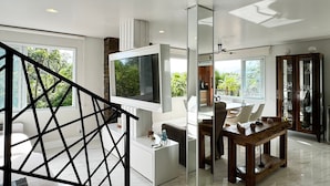 A modern living room with a large flat-screen TV, a dining area with a wooden table, and a staircase with a geometric railing. - Paraíso Azul do Rosa - IMB004 (Imbituba)