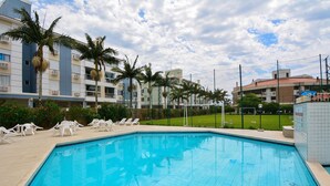 A swimming pool with lounge chairs and palm trees, surrounded by buildings and a grassy area. - Refúgio Terra do Sol - NIN009 (Florianópolis)