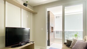 Un salon avec un téléviseur à écran plat posé sur un meuble en bois, un canapé avec des coussins, une petite plante sur une étagère et une porte-fenêtre coulissante donnant sur un balcon aménagé avec du mobilier d’extérieur.