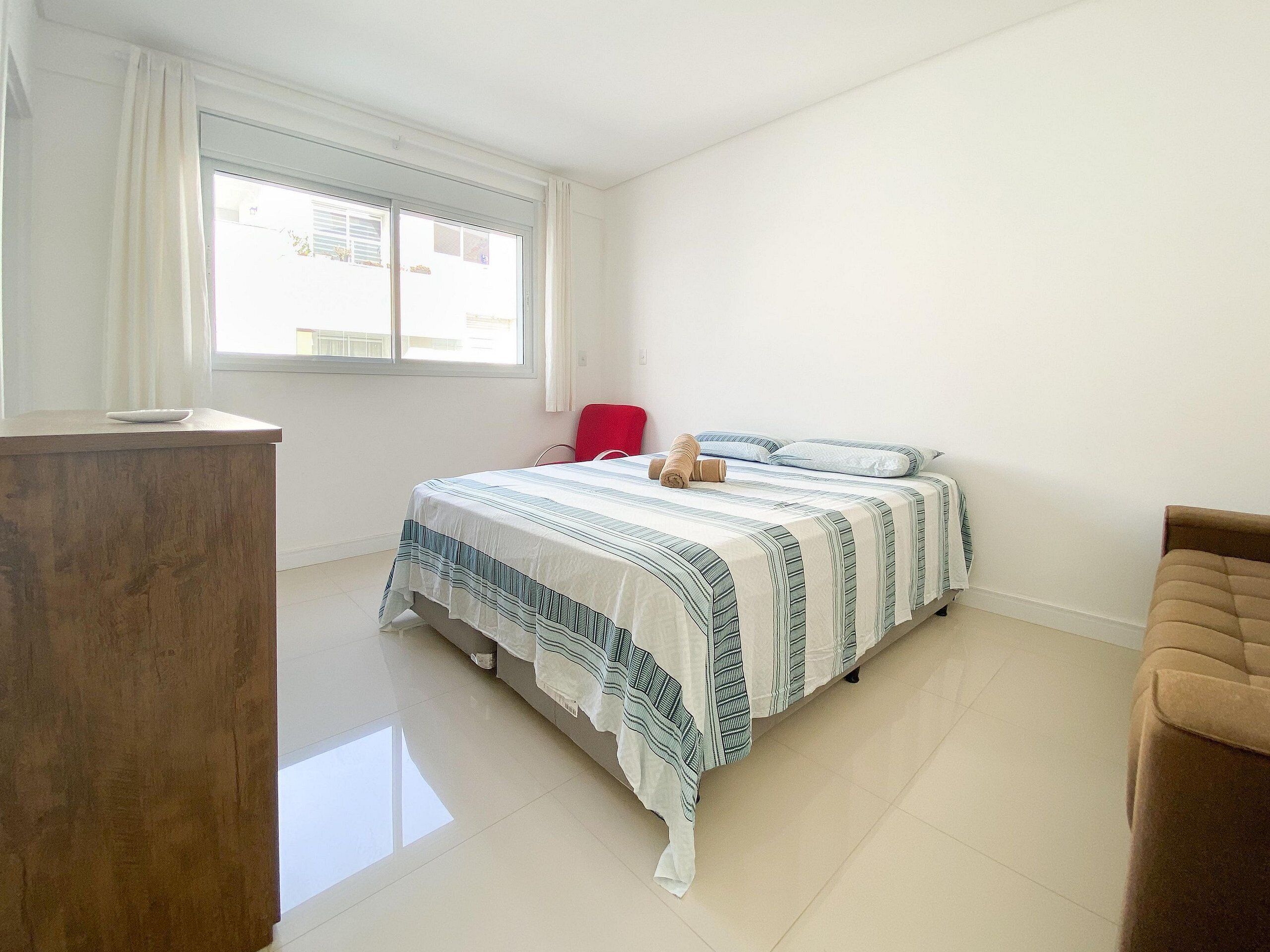 A bedroom with a bed, a red chair, a wooden desk, and a window with curtains.