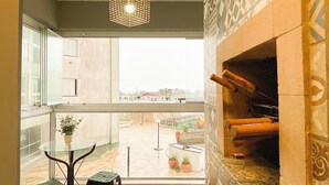 A balcony with a glass table and two stools, a fireplace, and a view of a pool and buildings. - Resid. Clube Mar Grosso - Laguna - LAG208 (Laguna)