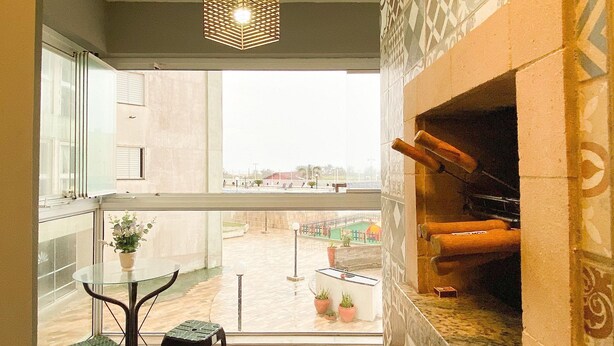 Un balcon avec une table en verre et deux tabourets, une cheminée, et une vue sur une piscine et des immeubles.