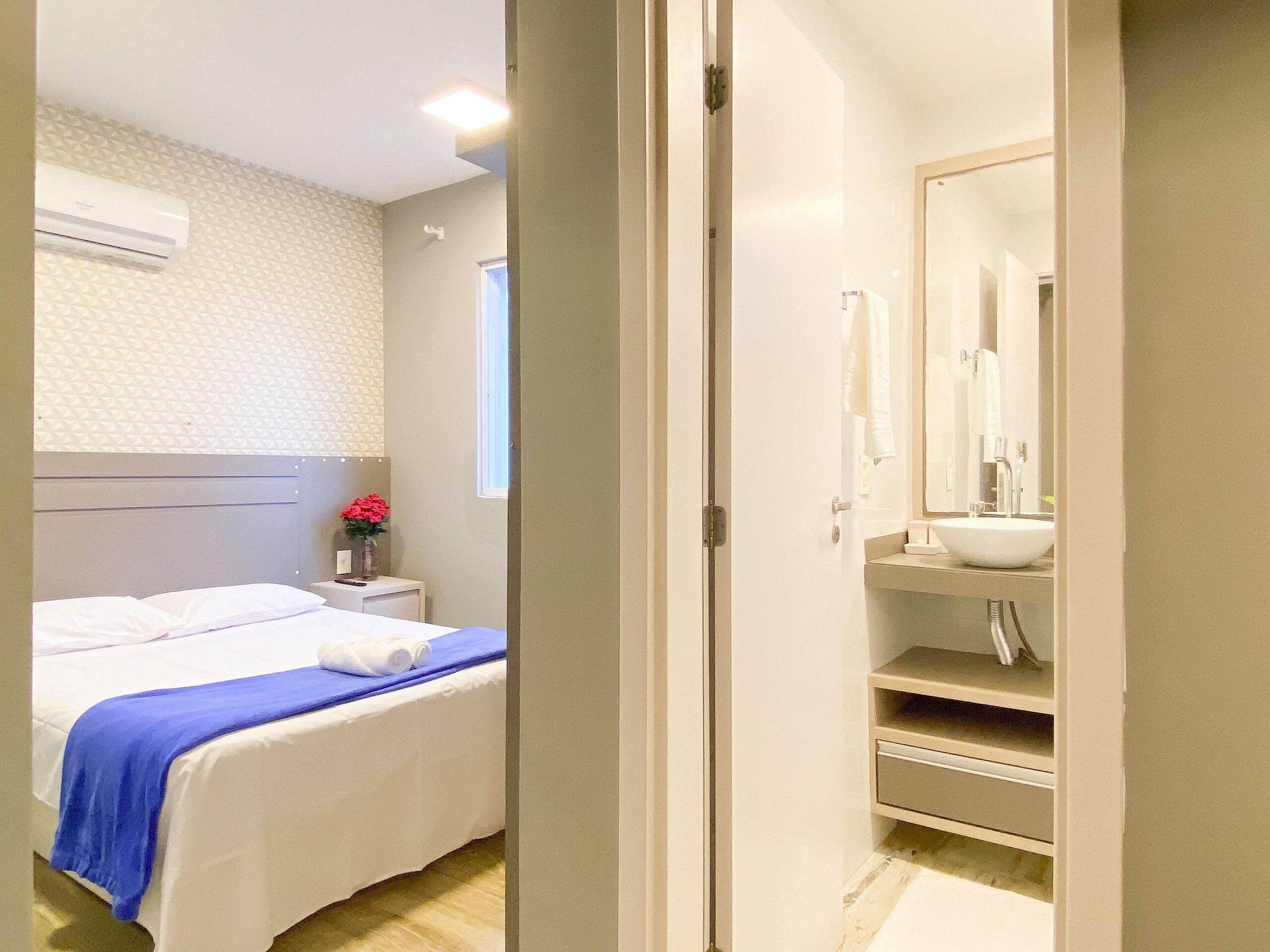 Une chambre d’hôtel comprenant un lit, une table de chevet avec un vase de fleurs, une salle de bain avec un lavabo et un miroir, et une porte donnant sur une autre pièce.