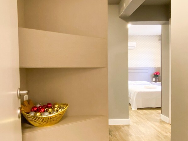 Una habitación de hotel con una cama, una mesita de noche con un florero y un pasillo que conduce a otra habitación.
