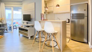 A modern kitchen with a built-in refrigerator, a dining area with two white chairs, a TV, and a coffee station. - Resid. Clube Mar Grosso - Laguna - LAG208 (Laguna)