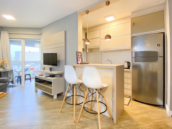 Una cocina moderna con refrigerador empotrado, una zona de comedor con dos sillas blancas, un televisor y una estación de café.