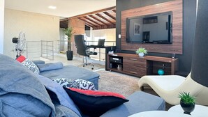 Ein modernes Wohnzimmer mit einem großen Flachbildfernseher an einer dunklen Wand, einem bequemen Sofa mit gemusterten Kissen, einem kleinen runden Tisch und einem Schreibtisch mit Stuhl und Topfpflanze.