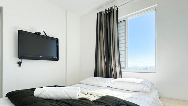 A hotel room with a bed, a flat-screen TV mounted on the wall, a window with curtains, and a view of the cityscape. - Resid. Clube Mar Grosso - Laguna - LAG801 (Laguna)