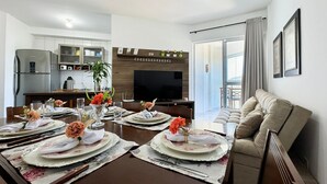 A dining area with a long table set for four, a kitchen in the background, and a balcony with curtains. - Resid. Clube Mar Grosso - Laguna - LAG801 (Laguna)