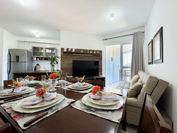 Un coin repas avec une longue table dressée pour quatre personnes, une cuisine en arrière-plan et un balcon avec des rideaux.