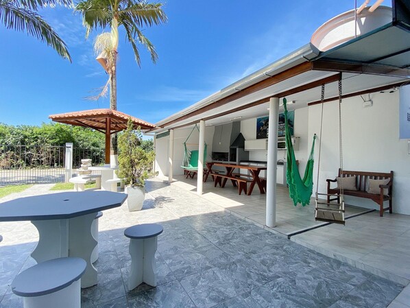 An outdoor patio area with a covered seating area, a dining table, and a swing chair. - Residencial Verdes Mares (Bombinhas)