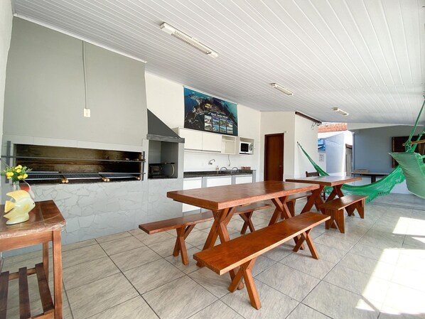 A kitchen with a wooden table and benches, a hanging hammock, and a poster on the wall. - Residencial Verdes Mares (Bombinhas)