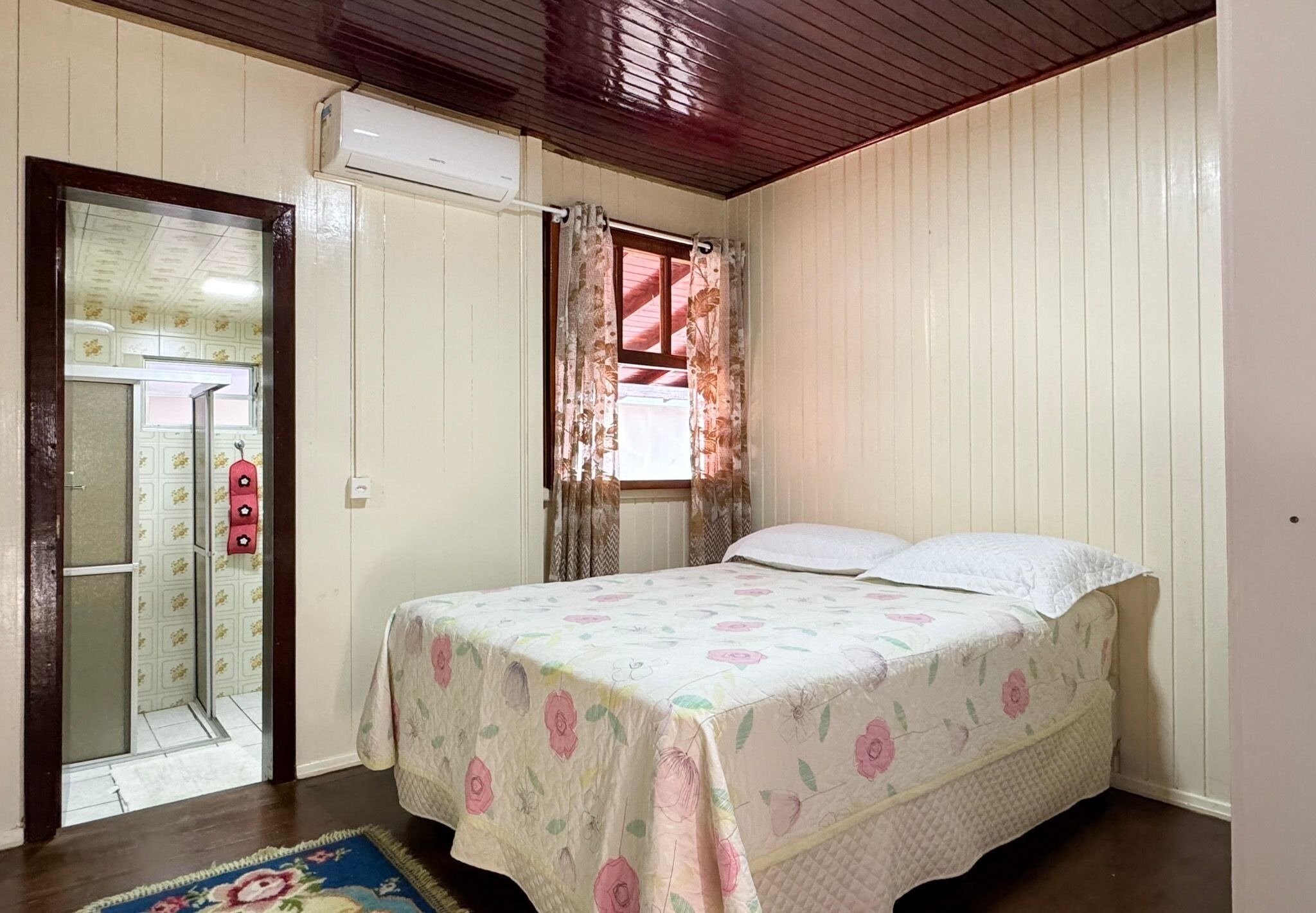 A bedroom with a bed, a window with curtains, a door leading to a bathroom, and a wooden ceiling.