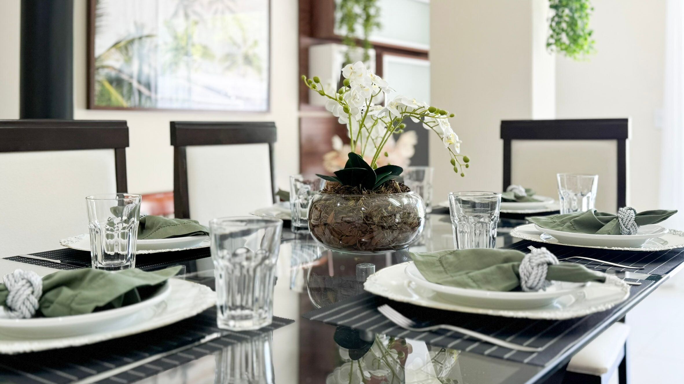Une table à manger dressée avec des assiettes, des verres et un centre de table composé de fleurs blanches dans un vase en verre.