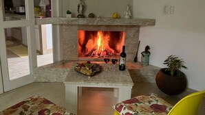 Una sala de estar con chimenea, una mesa de comedor con una bandeja de frutas y copas de vino, y dos sillas amarillas.