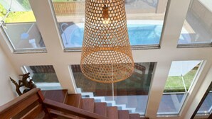 A modern interior with a wooden staircase, a hanging woven light fixture, and large windows offering a view of a swimming pool. - Casa de Andréa com piscina - Campeche - SDI004 (Florianópolis)