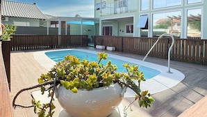 A pool area with a large potted plant in the foreground, wooden deck, and a two-story building with large windows in the background. - Casa de Andréa com piscina - Campeche - SDI004 (Florianópolis)
