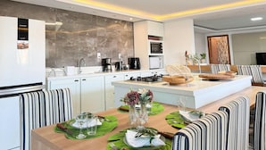 A modern kitchen with a central island, white cabinets, and a dining area with striped chairs and a wooden table set for a meal. - Casa de Andréa com piscina - Campeche - SDI004 (Florianópolis)