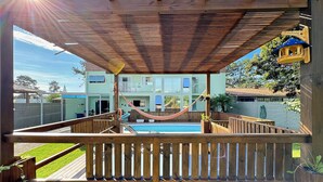 A wooden deck with a hammock, a pool, and a view of a two-story building. - Casa de Andréa com piscina - Campeche - SDI004 (Florianópolis)
