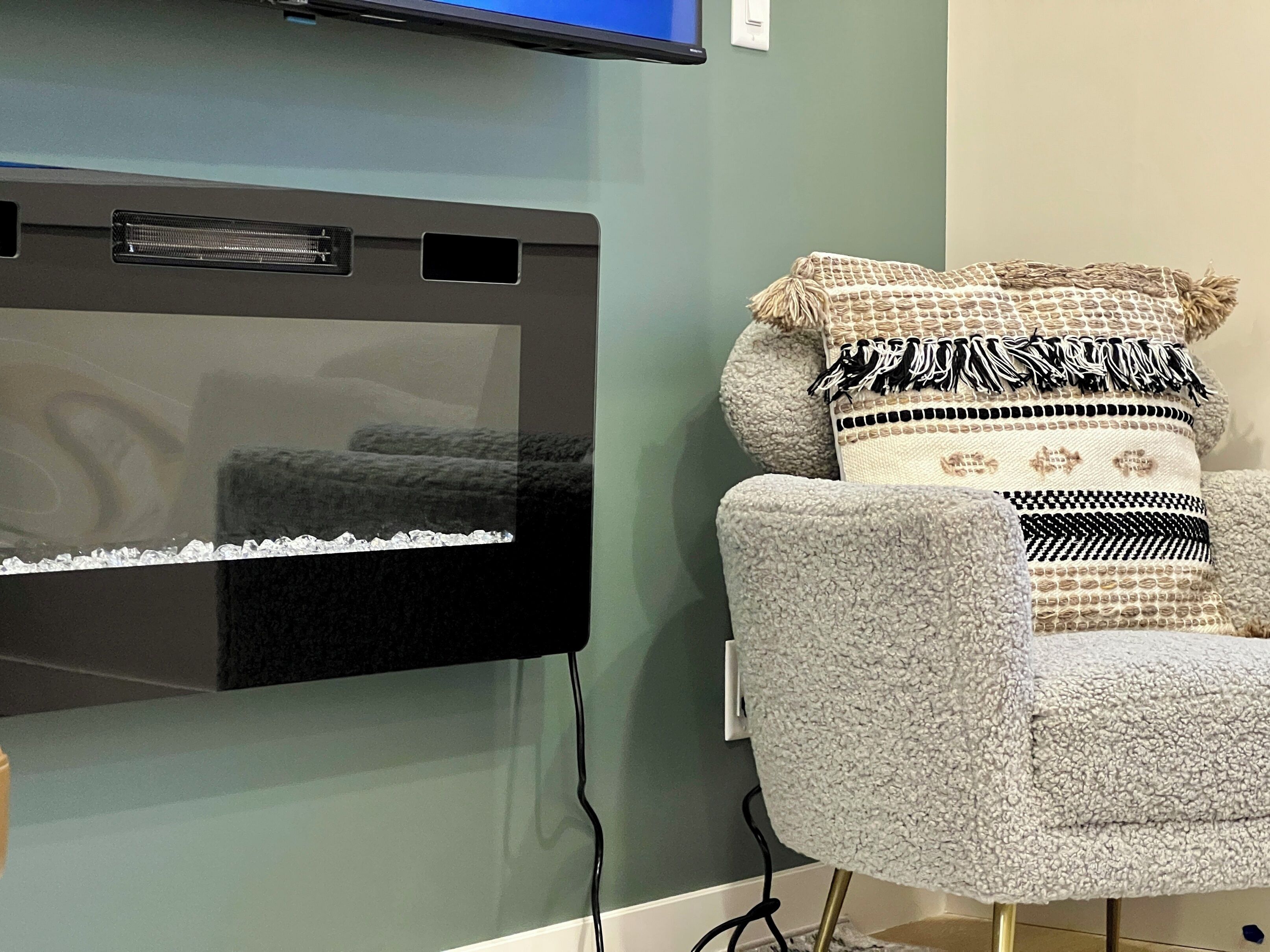Een kamer met een wandgemonteerde flatscreen-tv, een grijze fauteuil met gestreepte kussens en een stopcontact aan de muur.