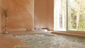 Een hottub met betegelde vloer, een decoratieve vaas en een raam met uitzicht op begroeiing.