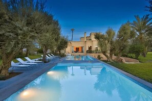 A swimming pool with lounge chairs, surrounded by trees, and a building with an orange-lit entrance. - Palais Rim Marrakech – Grande Villa de Vacances avec Piscine et Jardin (Marrakech)