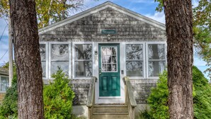 A small, two-story building with a green door and white windows, surrounded by trees and a stone wall.