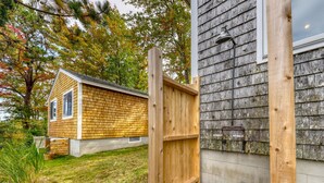 A wooden deck with a wooden fence, a small house with a shingled roof, and a concrete wall with a metal pipe.