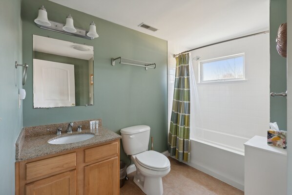 A bathroom with a white toilet, a sink with a granite countertop, wooden cabinets, a towel rack, a shower curtain, and a window.