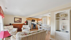 A living room with floral-patterned sofas, a dining table with chairs, a kitchen area with wooden cabinets, and a white cabinet with decorative items. - RARE 3 Bedroom at the Resort at Goose Rocks Beach (Kennebunkport)