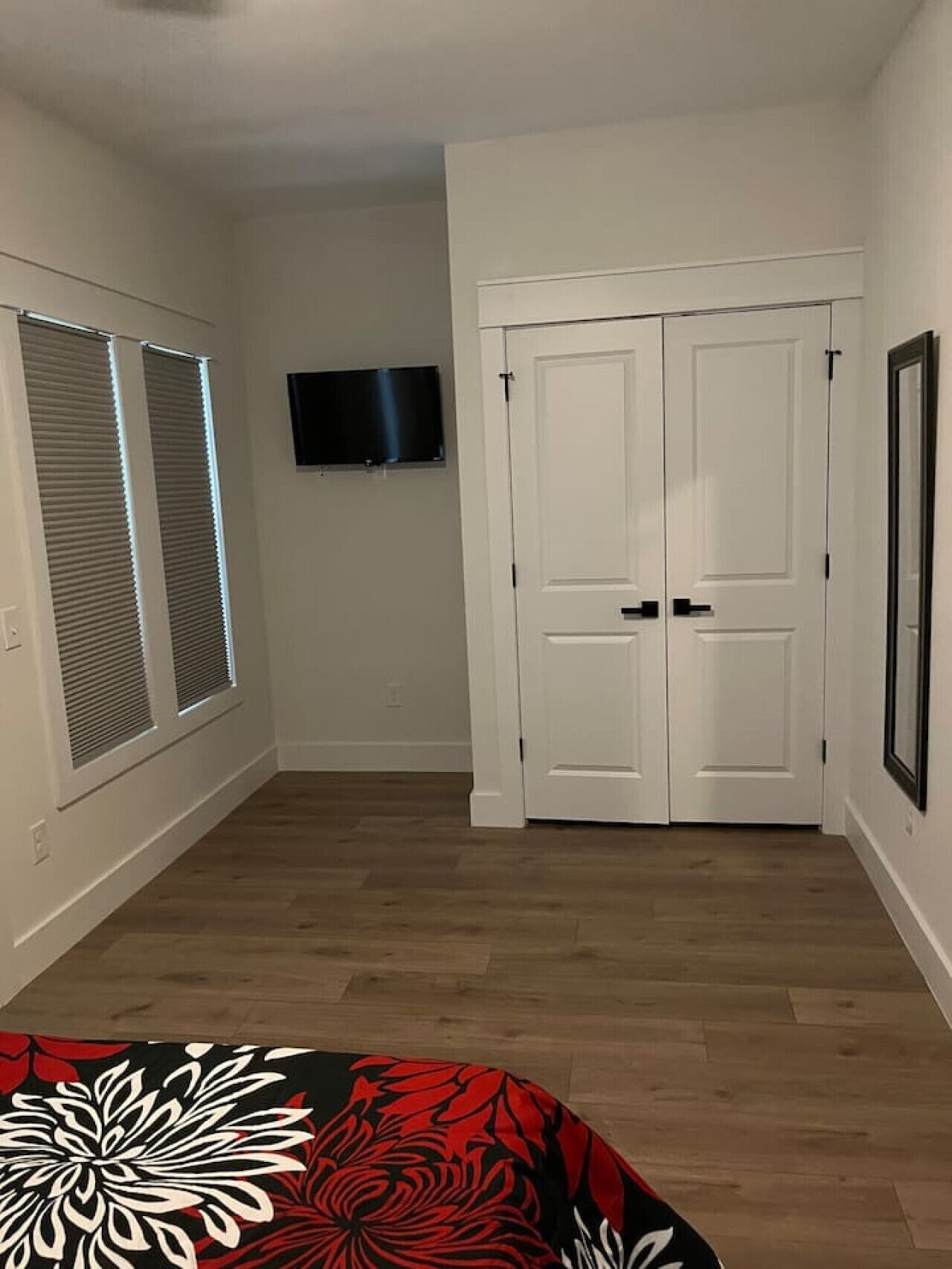 A room with a wooden floor, a bed with a floral bedspread, a TV mounted on the wall, and a set of white double doors.